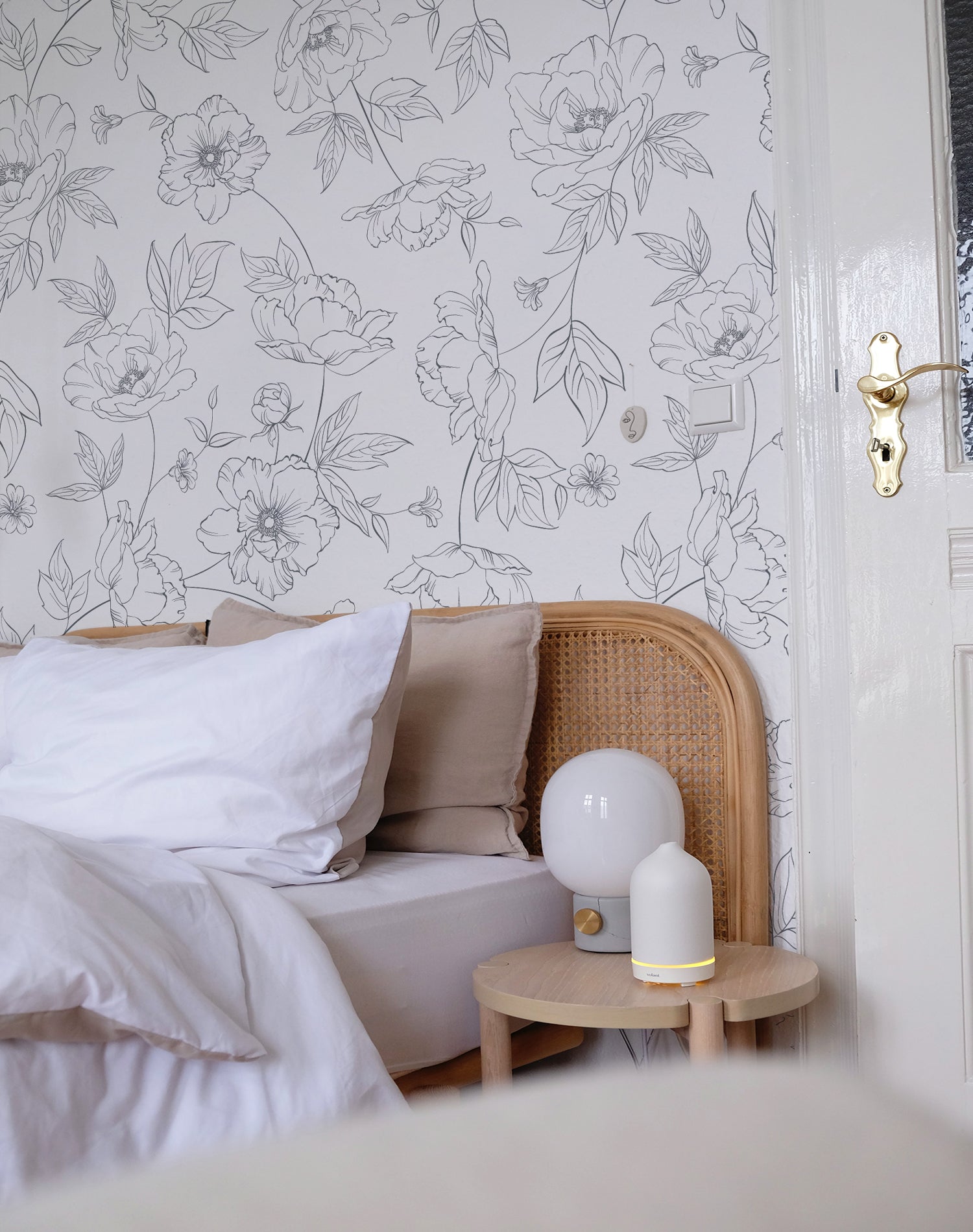 A stylish bedroom featuring a rattan headboard, white bedding, and a round wooden nightstand with a modern lamp. The wall behind the bed is covered with white wallpaper displaying a dainty black floral pattern, adding a touch of sophistication.