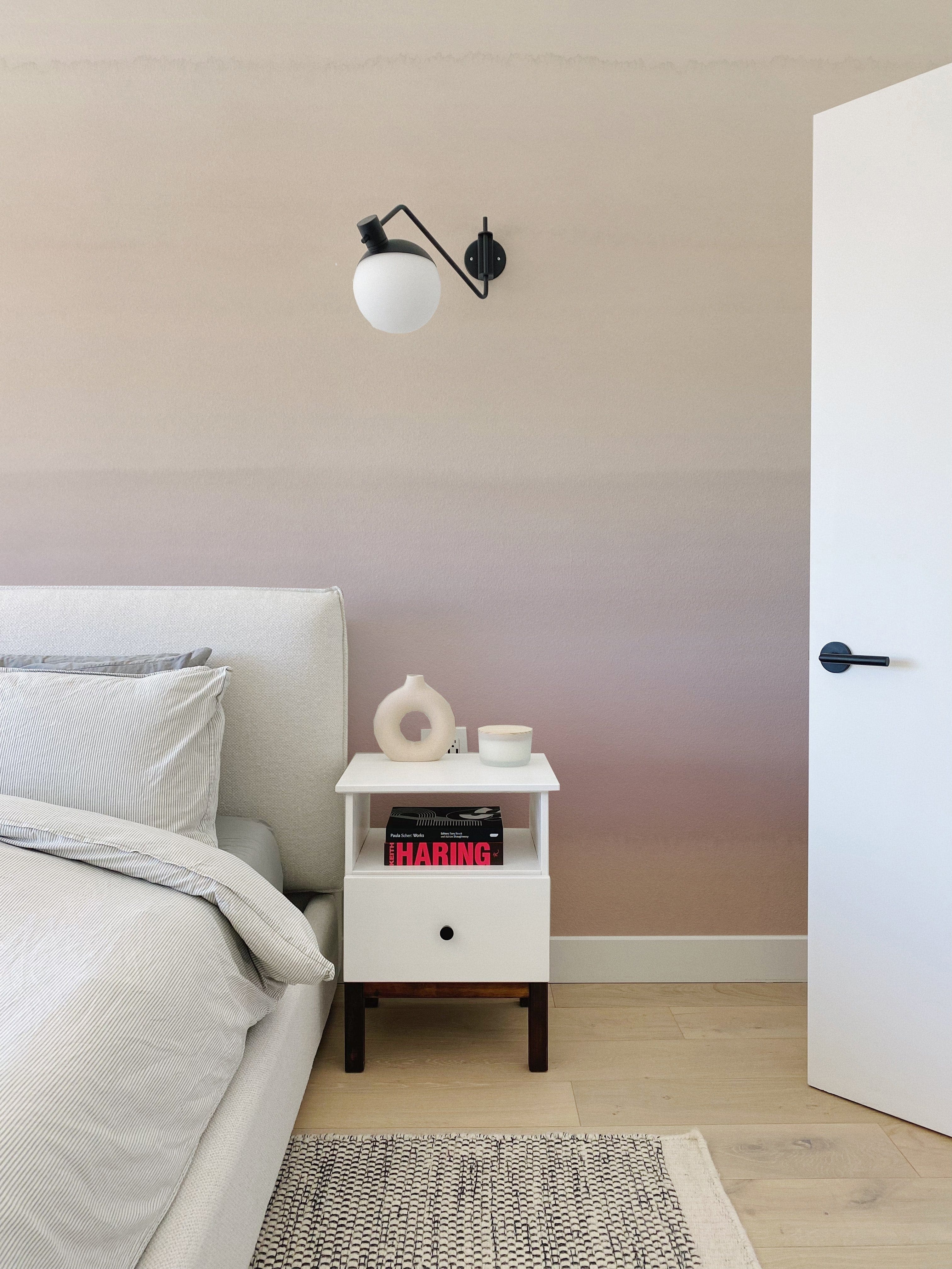 A modern bedroom featuring a wall covered in Blush Gradient Wallpaper with smooth transition from light beige at the top to a soft pink hue at the bottom. The scene is complemented by a white bedside table with books, a small vase, and a stylish black wall-mounted lamp.