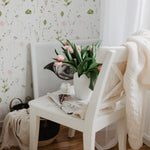 A charming and tranquil corner of a room featuring a chair with a white frame against a wall covered with Watercolour Floral Wallpaper in Blush Pink. The wallpaper depicts a beautiful array of watercolor flowers in soft pink, tender green leaves, and delicate brown branches, creating a gentle and romantic ambiance. A white vase on the chair holds a bouquet of fresh tulips, complemented by a cozy cream knit blanket draped over the chair's back and a decorative magazine, evoking a serene and inviting space.