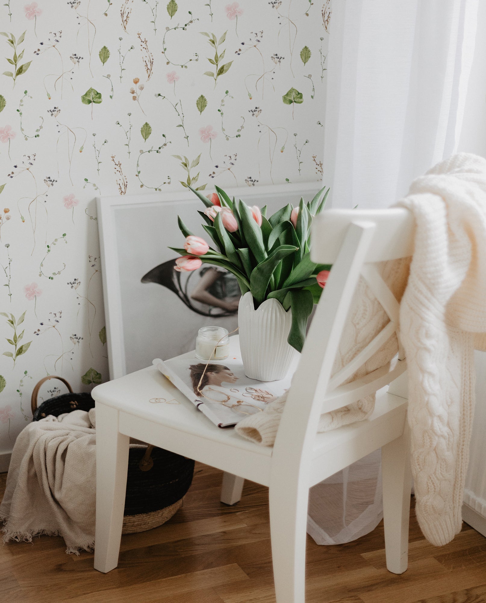 A charming and tranquil corner of a room featuring a chair with a white frame against a wall covered with Watercolour Floral Wallpaper in Blush Pink. The wallpaper depicts a beautiful array of watercolor flowers in soft pink, tender green leaves, and delicate brown branches, creating a gentle and romantic ambiance. A white vase on the chair holds a bouquet of fresh tulips, complemented by a cozy cream knit blanket draped over the chair's back and a decorative magazine, evoking a serene and inviting space.