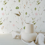 A minimalistic and serene composition against a wall adorned with the Watercolour Floral Wallpaper in Blush Pink. A uniquely shaped white vase sits on a white surface, holding a singular dried grass plume that gracefully leans to one side. The softness of the watercolor florals in the background pairs perfectly with the simplicity of the vase and the natural element of the grass, reflecting a modern and organic aesthetic