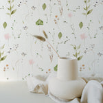 A minimalistic and serene composition against a wall adorned with the Watercolour Floral Wallpaper in Blush Pink. A uniquely shaped white vase sits on a white surface, holding a singular dried grass plume that gracefully leans to one side. The softness of the watercolor florals in the background pairs perfectly with the simplicity of the vase and the natural element of the grass, reflecting a modern and organic aesthetic.