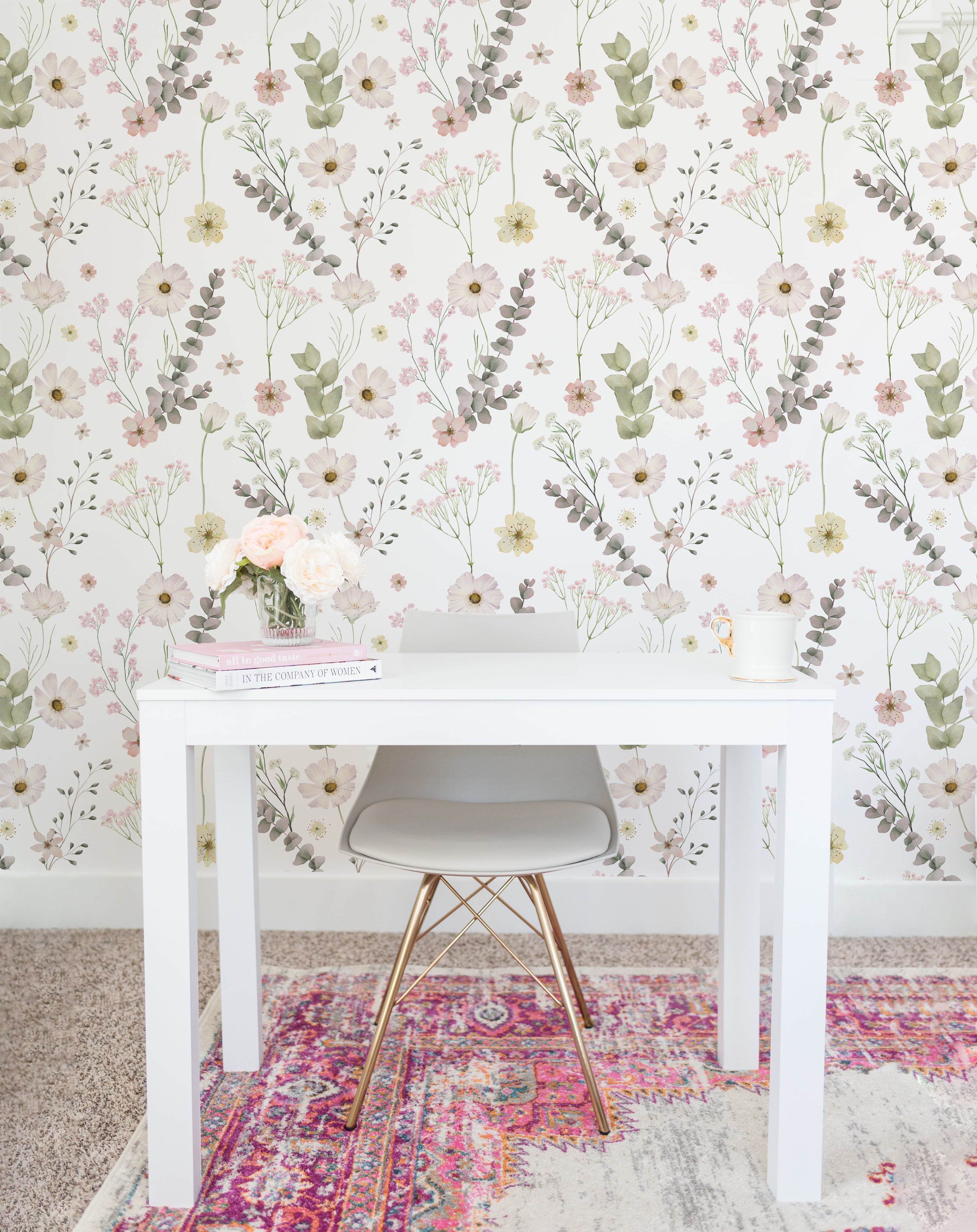A well-lit home office corner with a white desk against a wall adorned with the Botanical Muse Wallpaper, featuring a pattern of soft-colored wildflowers and eucalyptus leaves. On the desk, a gray modern chair with gold legs, a small vase of peach roses, books, and a candle create a serene workspace.