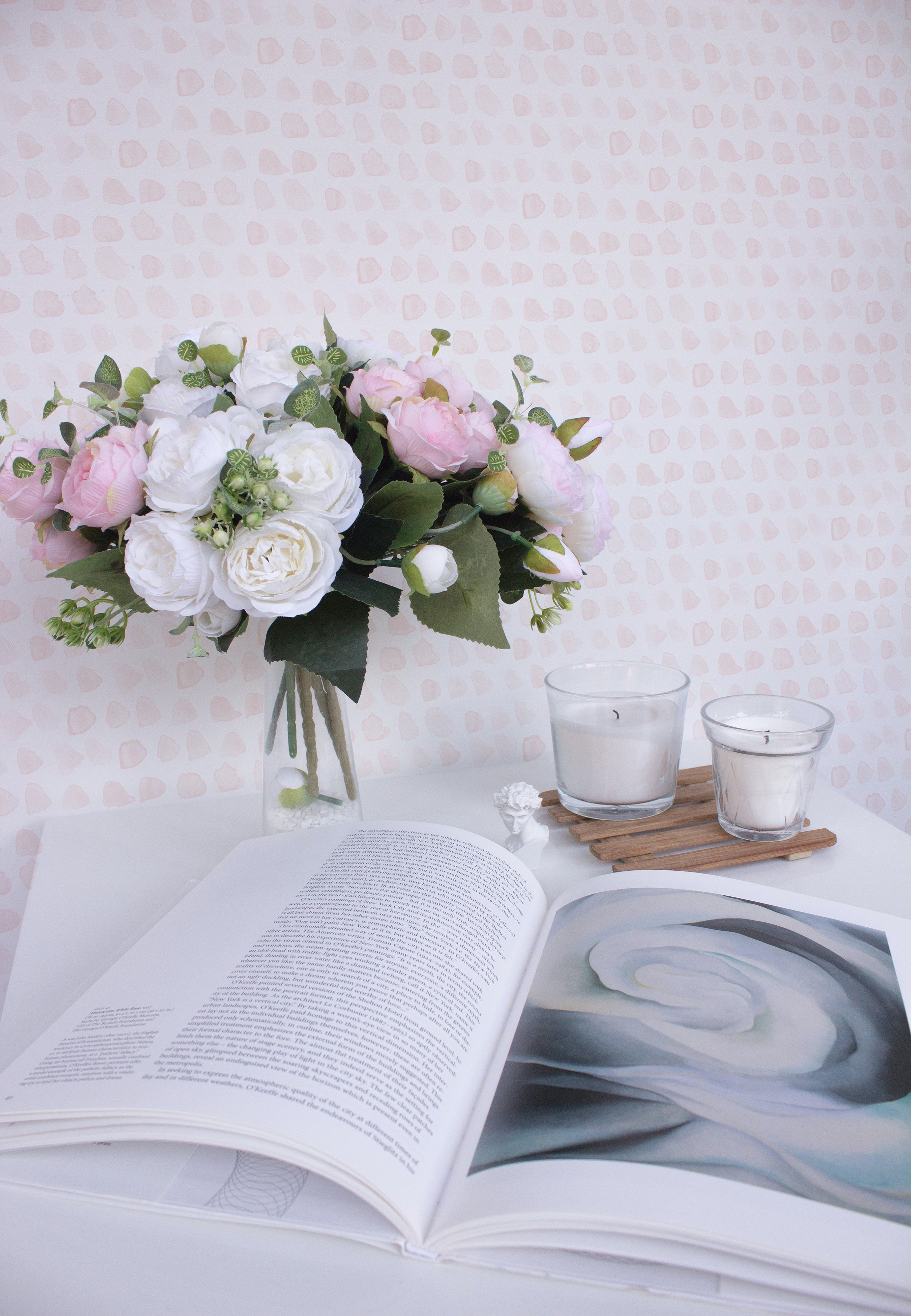 A serene setting featuring the Handpainted Dots Wallpaper as a backdrop. A vase filled with an arrangement of lush white and pink flowers is placed to the left, with a wooden tray holding two white candles in glass holders below. An open book with visible text lies in the foreground, showcasing an artistic image on the right page.