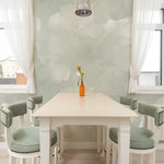 A dining room with a white table and chairs with sage green upholstery, placed against a wall covered with light sage cloud mural wallpaper, featuring soft, cloudy patterns.