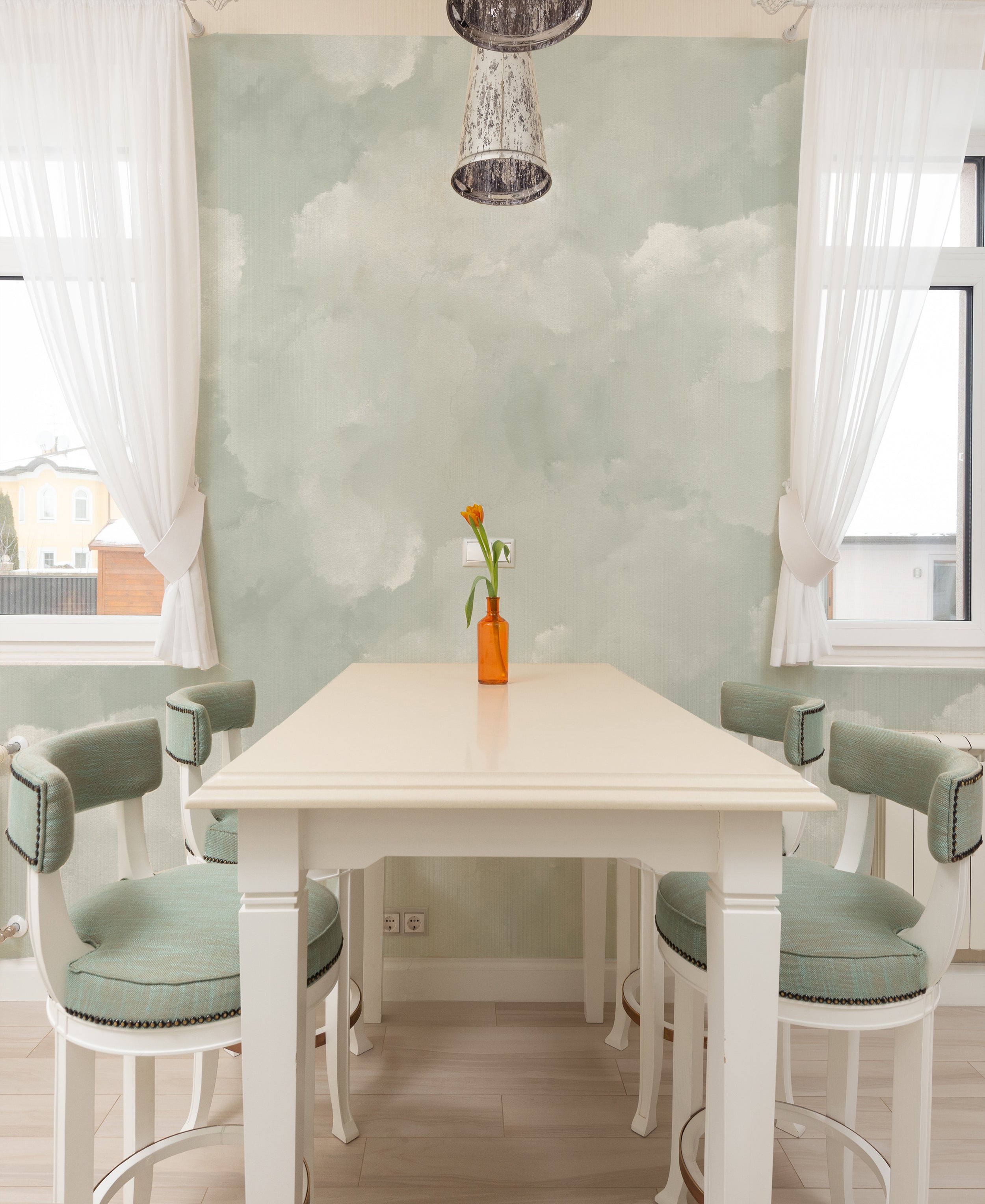 A dining room with a white table and chairs with sage green upholstery, placed against a wall covered with light sage cloud mural wallpaper, featuring soft, cloudy patterns.