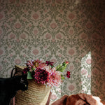 A cozy corner with a woven basket filled with lush pink flowers sits beside a black cat and a crumpled terracotta-colored blanket, against a backdrop of pastel floral damask wallpaper.
