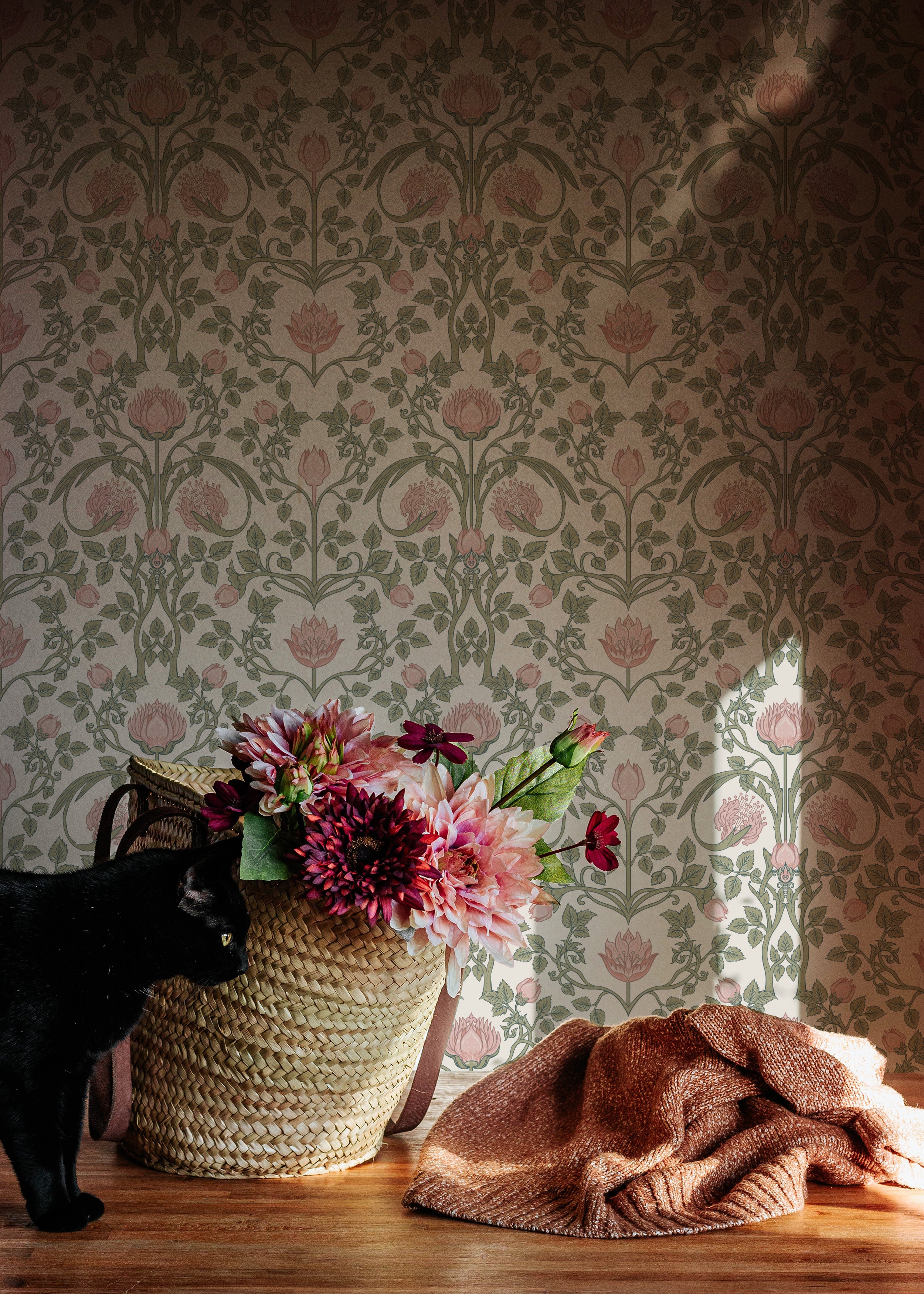 A cozy corner with a woven basket filled with lush pink flowers sits beside a black cat and a crumpled terracotta-colored blanket, against a backdrop of pastel floral damask wallpaper.
