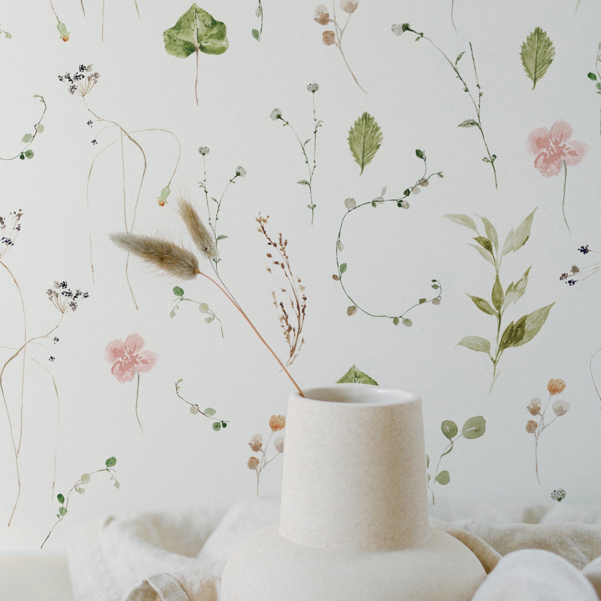 A serene and artistic display featuring the Modern Watercolour Floral Wallpaper in Dusty Pink as a backdrop. The wallpaper showcases a delicate array of watercolor flowers and foliage in shades of pink, green, and beige, giving the impression of a hand-painted wall. A sculptural vase in front of the wallpaper adds to the soft and organic feel of the setting.