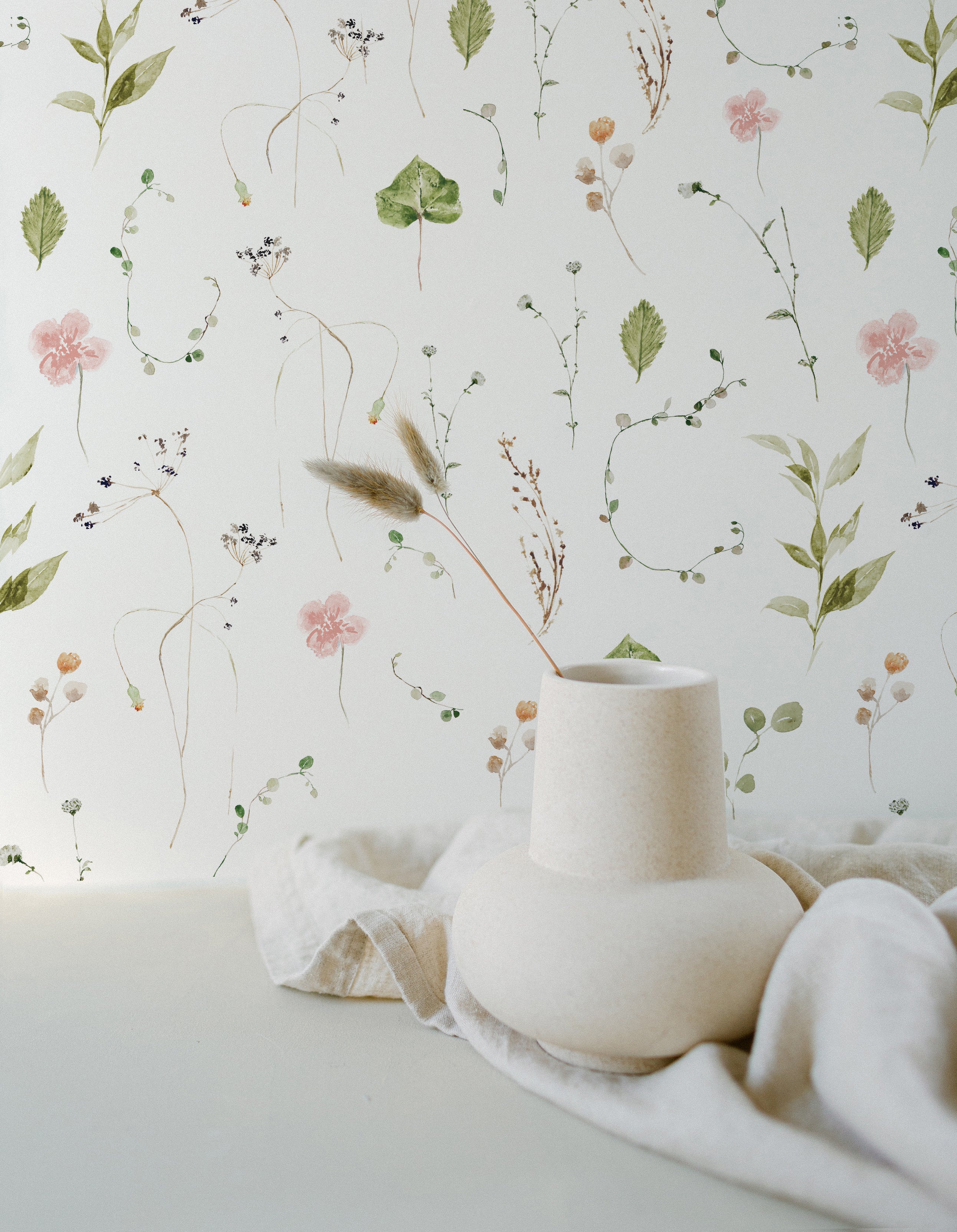 A serene and artistic display featuring the Modern Watercolour Floral Wallpaper in Dusty Pink as a backdrop. The wallpaper showcases a delicate array of watercolor flowers and foliage in shades of pink, green, and beige, giving the impression of a hand-painted wall. A sculptural vase in front of the wallpaper adds to the soft and organic feel of the setting.
