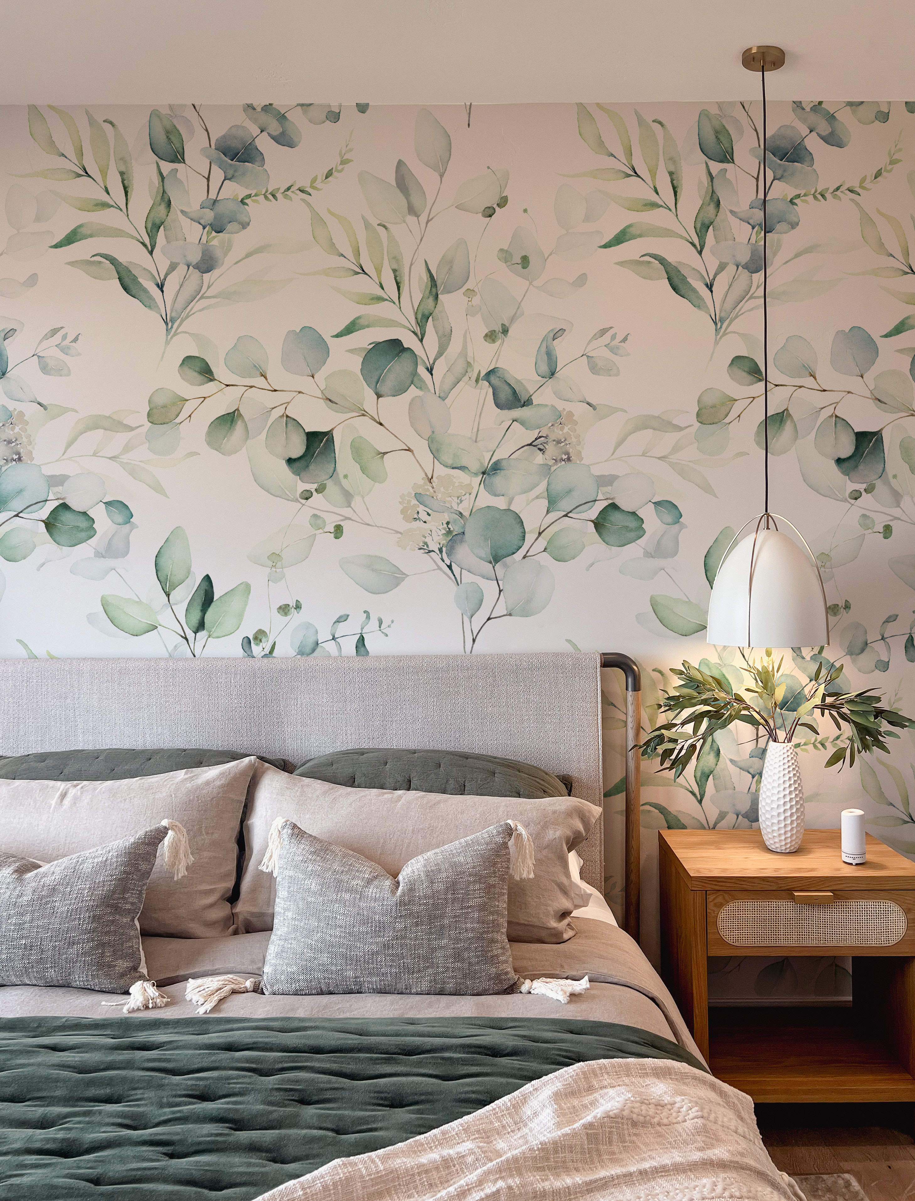 A stylish bedroom featuring the Green Leaves and Branches Wallpaper - Large, with an elegant botanical design of green leaves and branches that brings a sense of nature's serenity to the room, complemented by textured gray bedding and a chic bedside table with a vase and lamp.