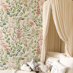 A whimsical children's room corner featuring a canopy bed with sheer drapery. The backdrop is adorned with Watercolour Floral and Leaf Wallpaper, a lush display of botanical illustrations with pink foxgloves, blueberries, and soft white flowers, all set against a creamy background. Assorted pillows and a plush toy create a comforting and dreamy sleep space