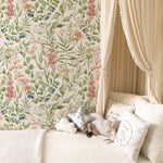 A whimsical children's room corner featuring a canopy bed with sheer drapery. The backdrop is adorned with Watercolour Floral and Leaf Wallpaper, a lush display of botanical illustrations with pink foxgloves, blueberries, and soft white flowers, all set against a creamy background. Assorted pillows and a plush toy create a comforting and dreamy sleep space