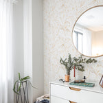 A chic and contemporary room setting featuring the Gold Tropical Wallpaper, with gold-leaf tropical plant illustrations adorning the wall, complemented by modern furnishings including a white dresser with gold handles, a brown leather pouf, and a round mirror.