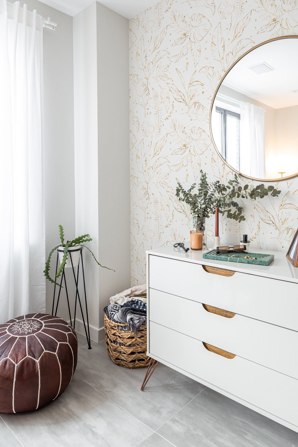 A chic and contemporary room setting featuring the Gold Tropical Wallpaper, with gold-leaf tropical plant illustrations adorning the wall, complemented by modern furnishings including a white dresser with gold handles, a brown leather pouf, and a round mirror.