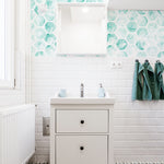 A modern bathroom featuring a vibrant turquoise hexagonal honeycomb wallpaper with a watercolor effect. The wallpaper adds a splash of color against the white subway tiles, complementing the minimalist white sink and turquoise towels.