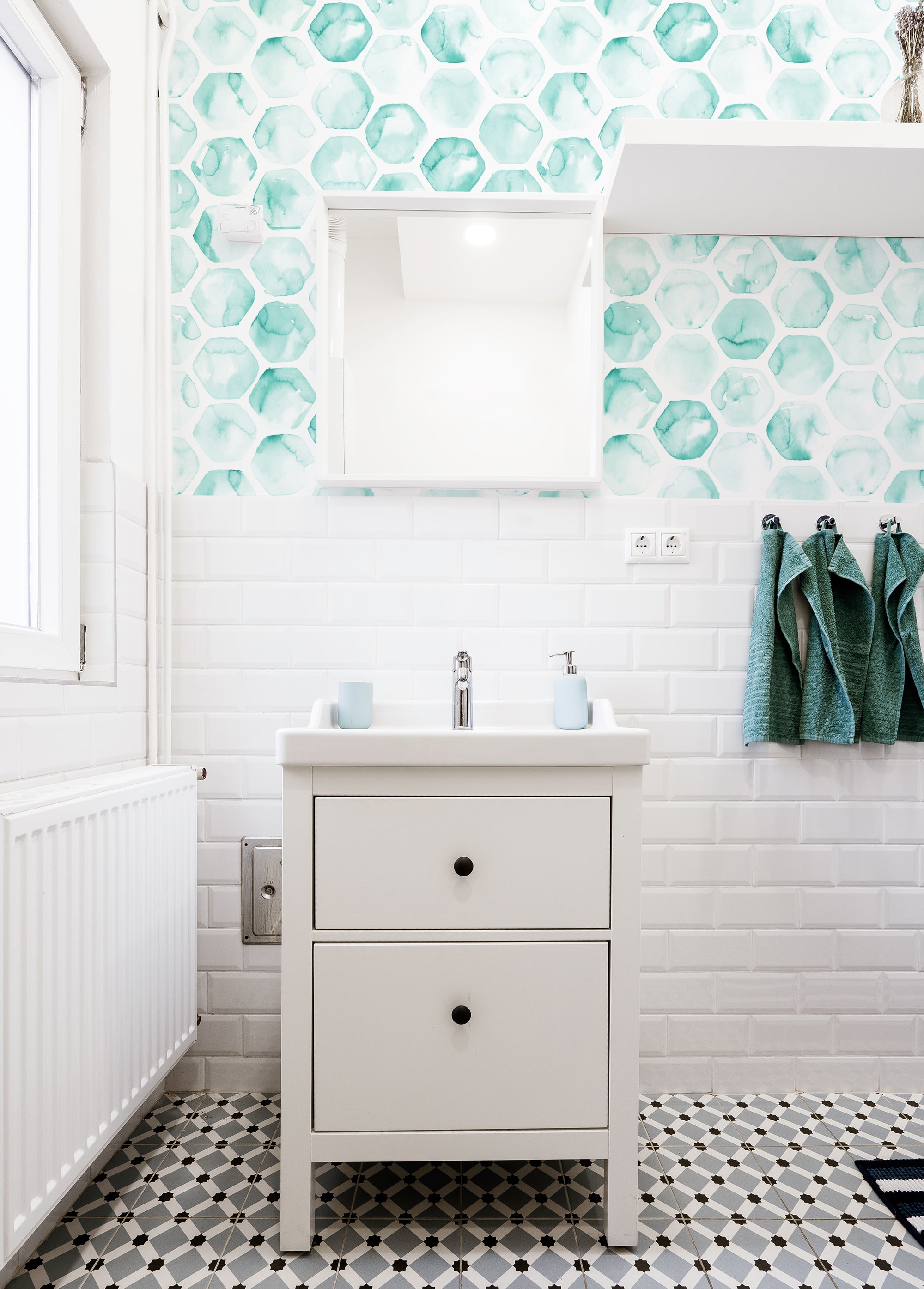 A modern bathroom featuring a vibrant turquoise hexagonal honeycomb wallpaper with a watercolor effect. The wallpaper adds a splash of color against the white subway tiles, complementing the minimalist white sink and turquoise towels.