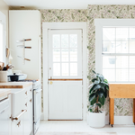 A cozy kitchen corner with vintage white cabinetry and a wooden countertop. A white door with a glass top half allows natural light to filter in, casting a warm glow on the floral wallpaper that lines the upper part of the walls, showcasing an array of small wildflowers.