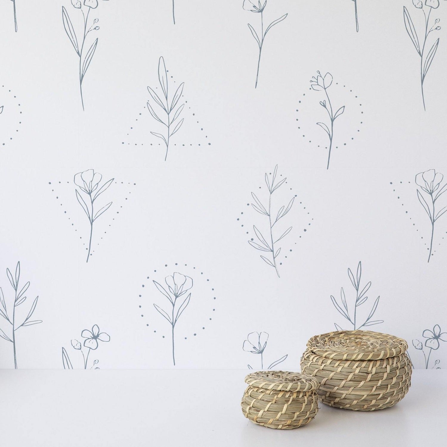 An interior setting with a segment of wall covered in the Minimal Floral Line Art Wallpaper. The pattern displays a series of delicate botanical illustrations in blue line art on a white background. In the foreground, two small woven baskets rest on a surface, complementing the natural theme.