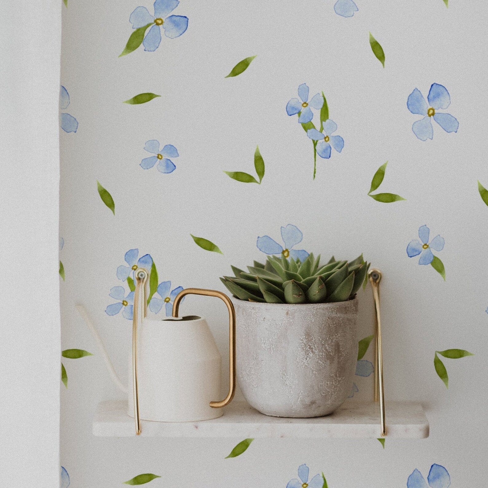 A stylish home corner featuring the Tiny Blue Watercolour Floral Wallpaper on the wall, with a decorative shelf holding a watering can and a succulent in a rustic pot. The soft floral pattern complements the minimalist and natural decor.