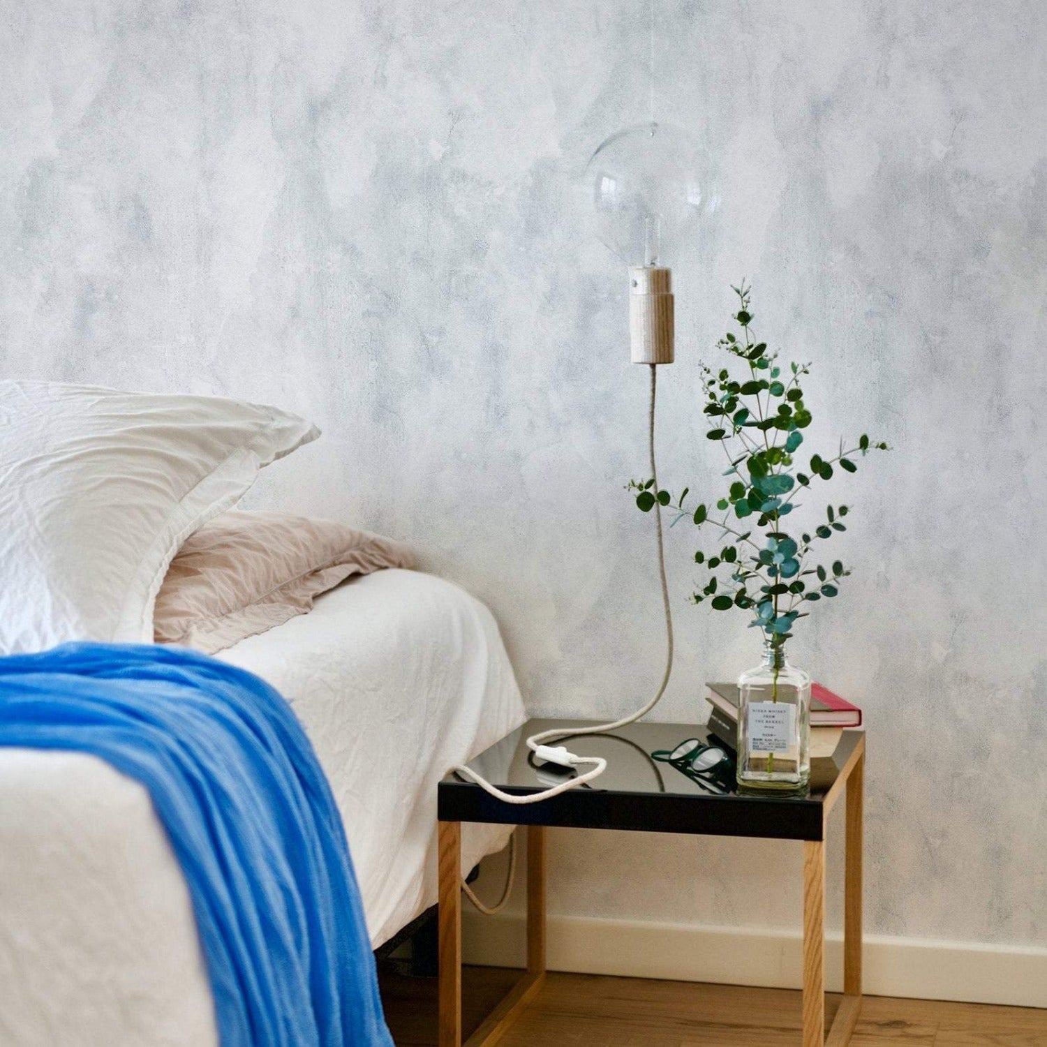 Bedroom interior featuring Blue Limewash Texture Wallpaper with a subtle, cloudy texture. The room includes a bed with white and beige bedding, a bright blue throw, a bedside table with a vase of greenery, and a modern pendant light, creating a tranquil and stylish ambiance.