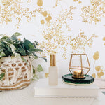 A stylish corner of a room featuring a wall adorned with Gold Floral Bunches Wallpaper, complemented by a woven planter with lush greenery and chic gold and glass decor accents.