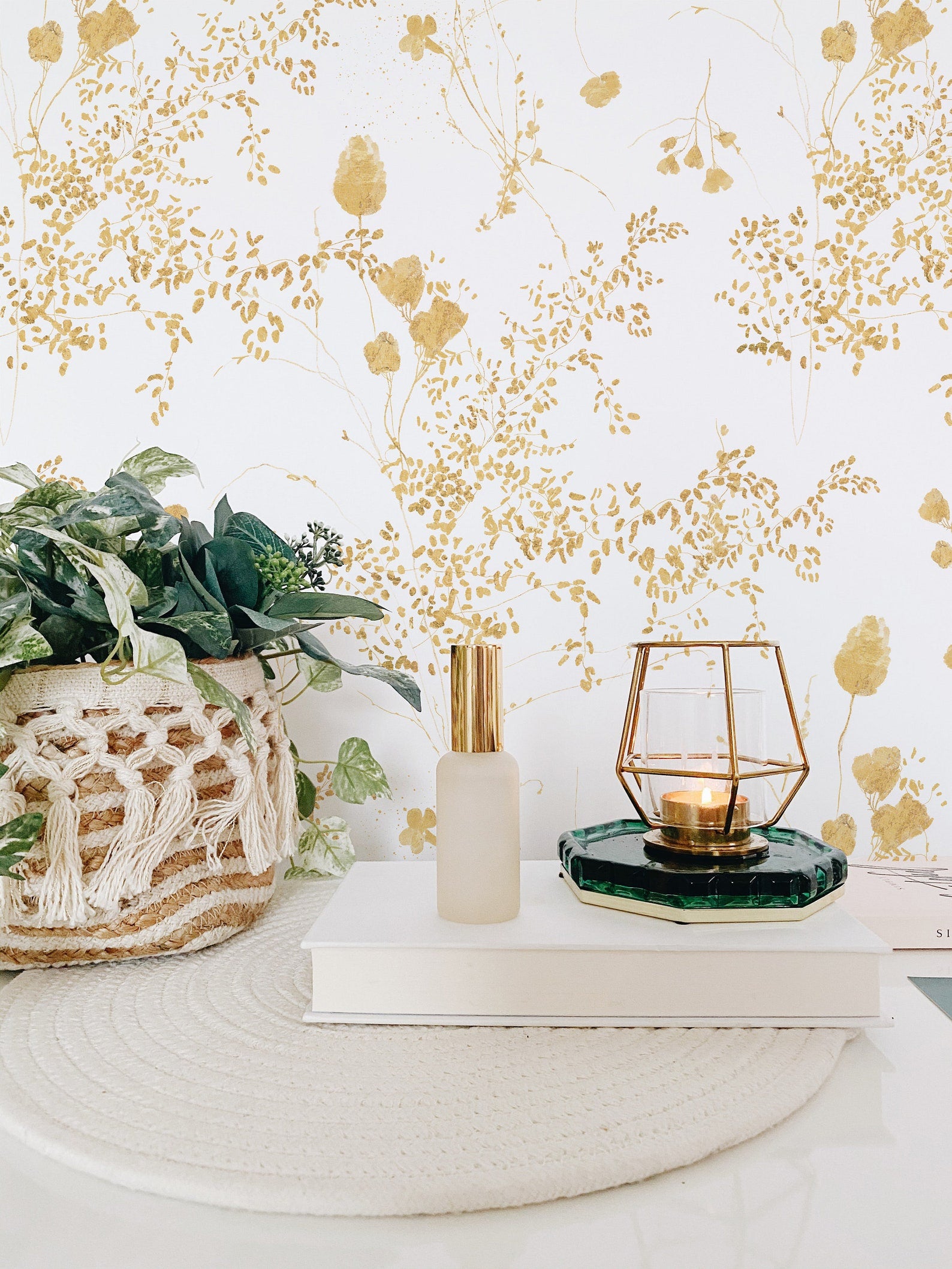 A stylish corner of a room featuring a wall adorned with Gold Floral Bunches Wallpaper, complemented by a woven planter with lush greenery and chic gold and glass decor accents.