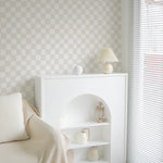 A tranquil corner of a room featuring the Funky Checkered Wallpaper in subtle beige tones, complemented by a cream armchair draped with a beige throw. A minimalist white shelf displays an assortment of neutral-toned decorative items, alongside a simple lamp, all bathed in natural light from the window.