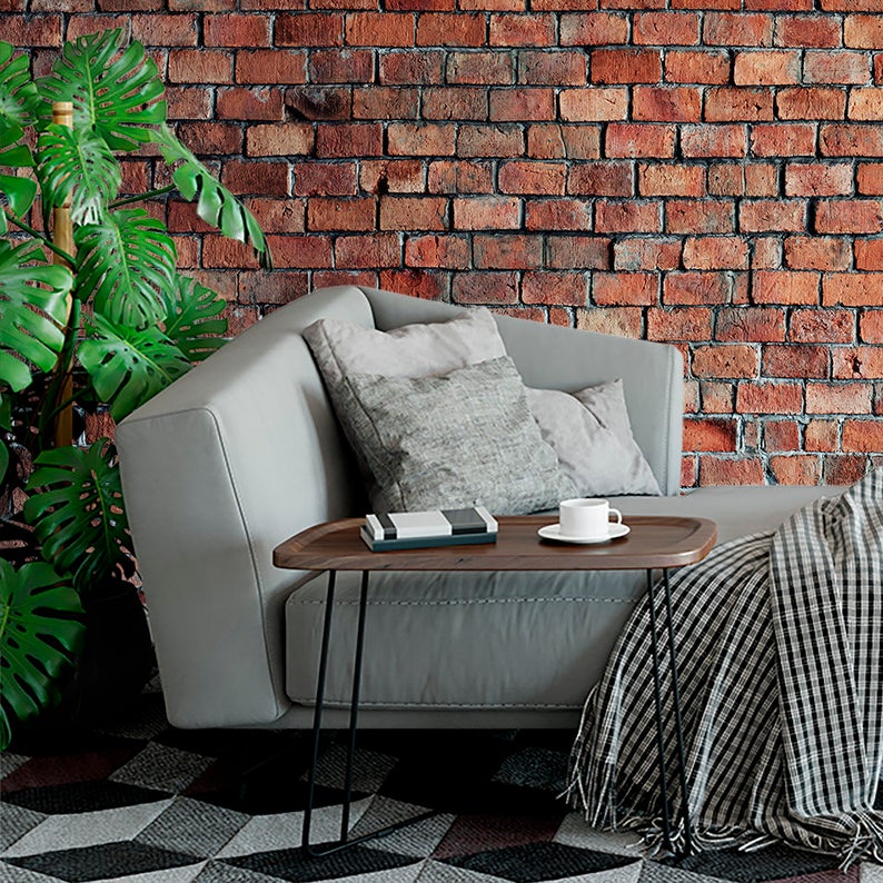 A modern living room corner with a realistic red brick wallpaper providing a rustic and textured background. A comfortable grey sofa with a plaid throw and a round coffee table with a couple of cups on top complement the urban loft atmosphere.