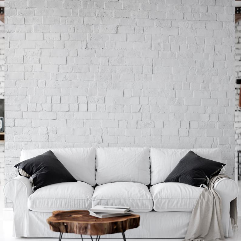 A clean and inviting living room featuring a white sofa with black throw pillows. The focal point of the room is the Realistic White Brick Wallpaper that gives the space a modern, urban feel, with its textured appearance lending depth and character to the wall behind the sofa. A rustic wooden coffee table in front of the sofa adds warmth to the decor.
