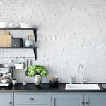 A modern kitchen setup showcasing the Realistic White Brick Wallpaper which serves as a stylish backsplash, complementing the grey cabinetry. The kitchen is well-appointed with contemporary appliances, open shelving with neatly arranged dishware, and a lush green plant in a vintage pot, adding a pop of color and life to the space.