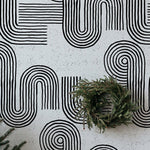 The Zen Abstract Wallpaper adds a touch of sophistication with its black and white abstract line patterns, reminiscent of a zen garden, against a speckled grey background, complemented by the simplicity of a pine wreath on the floor.