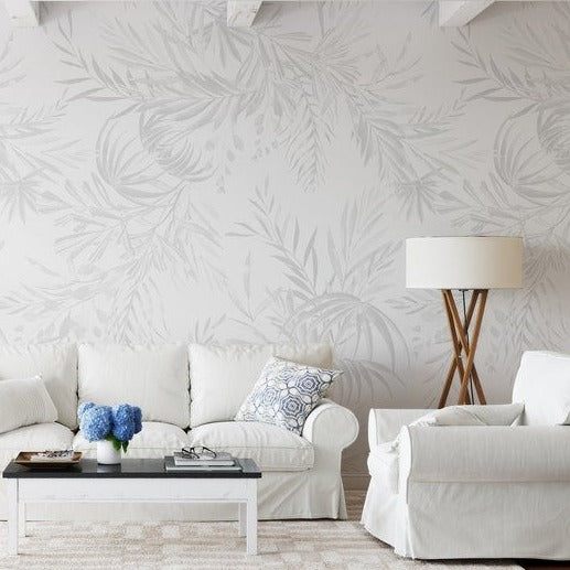 An elegant living room setting showcases the Palm Springs Wallpaper, which features a large-scale, soft grey palm leaf design, adding a touch of subtle sophistication to the decor. The wallpaper creates a serene backdrop for the white upholstered furniture and pops of blue decor accents.