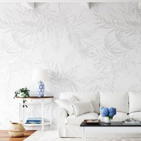 An elegant living room setting showcases the Palm Springs Wallpaper, which features a large-scale, soft grey palm leaf design, adding a touch of subtle sophistication to the decor. The wallpaper creates a serene backdrop for the white upholstered furniture and pops of blue decor accents.