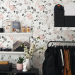 An interior wall covered with a pastel floral wallpaper, featuring delicate pink flowers and gray leaves, complemented by decorative shelves holding books, cameras, and a vase with white flowers, creating a cozy and stylish atmosphere