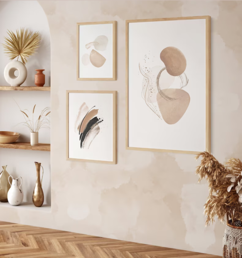 A home interior with abstract watercolor artwork displayed on the wall. Three framed pieces of art exhibit fluid shapes in neutral tones of beige, cream, and black. The wall is adorned with an organic watercolor mural in a matching color palette. Below the artwork, a wooden shelf holds decorative vases in complementary earthy tones