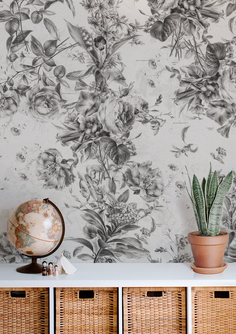 A cozy corner showcasing the Toile De Jouy Monochrome Floral Wallpaper, with detailed black and white botanical drawings, complemented by a vintage globe and potted snake plant atop a woven storage unit