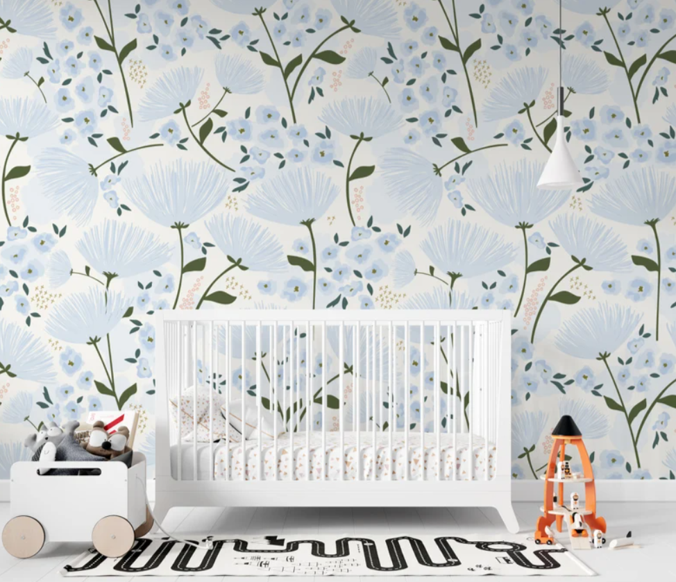 A serene nursery features the "Floral Love Wallpaper" with its beautiful blue floral pattern, creating a peaceful backdrop for a white crib. The room's decor is minimal and tasteful, allowing the intricate design of the wallpaper to stand out and fill the space with a calming botanical charm.