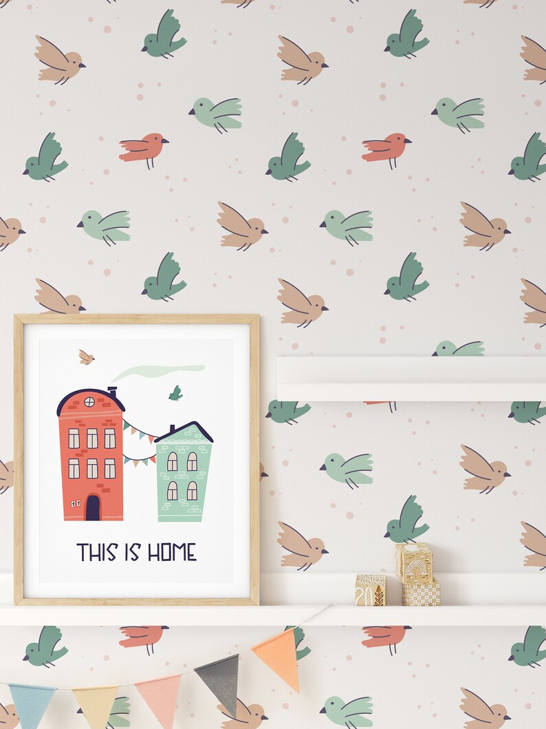 Framed art of two colorful houses with 'This Is Home' text against bird-patterned wallpaper in a nursery.