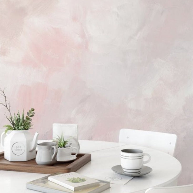 A chic dining area with a soft, Venetian watercolor wallpaper in hues of blush and cream creating an abstract backdrop. A round white table is set with tea-time accessories, complementing the wall’s dreamy aesthetic.