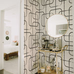 A stylish corner of a room with Midnight Wallpaper, featuring bold black lines creating a geometric grid pattern on a white background. A hanging wooden shelf displays home decor, and a canvas tote with abstract face embroidery leans against the wall.