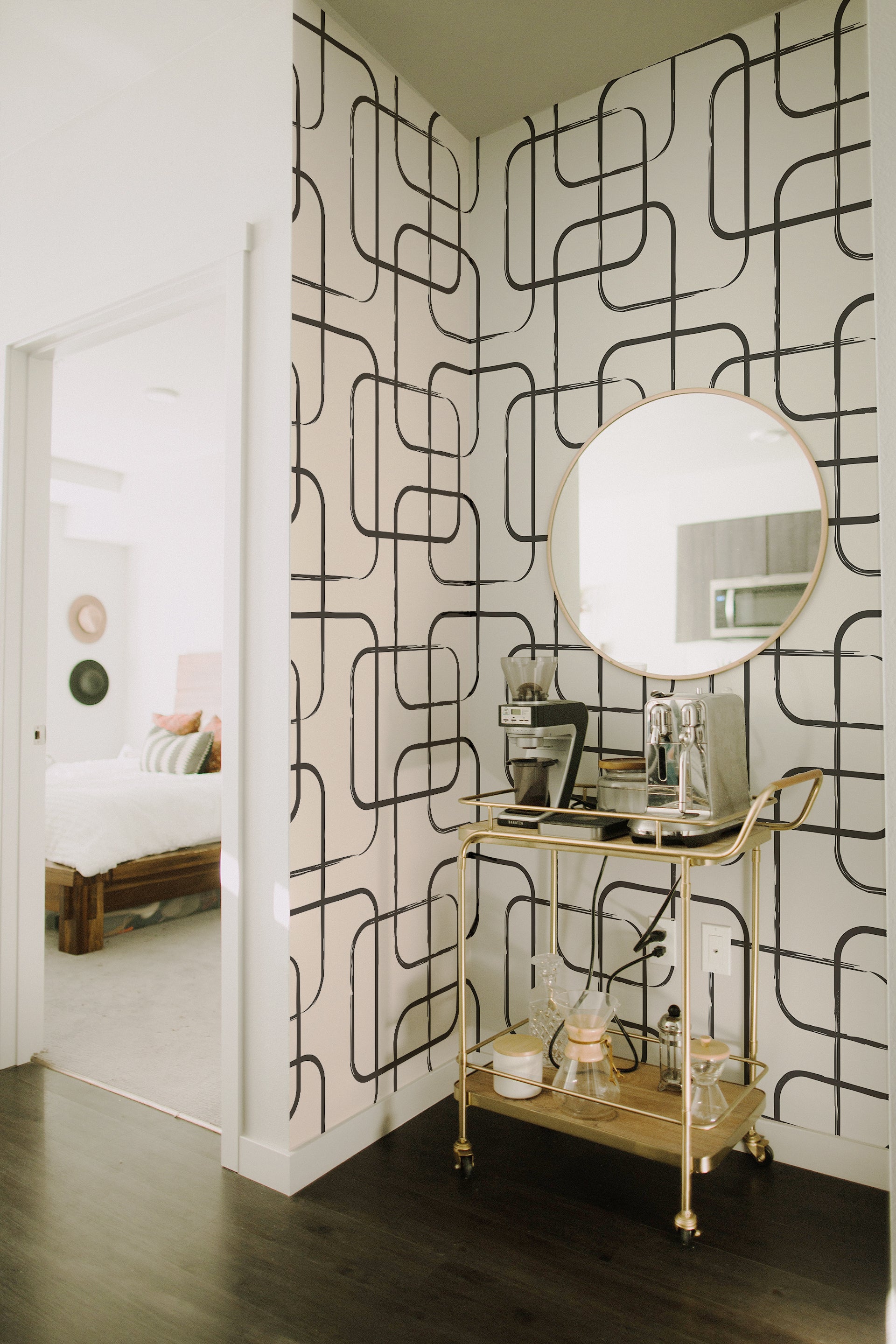 A stylish corner of a room with Midnight Wallpaper, featuring bold black lines creating a geometric grid pattern on a white background. A hanging wooden shelf displays home decor, and a canvas tote with abstract face embroidery leans against the wall.