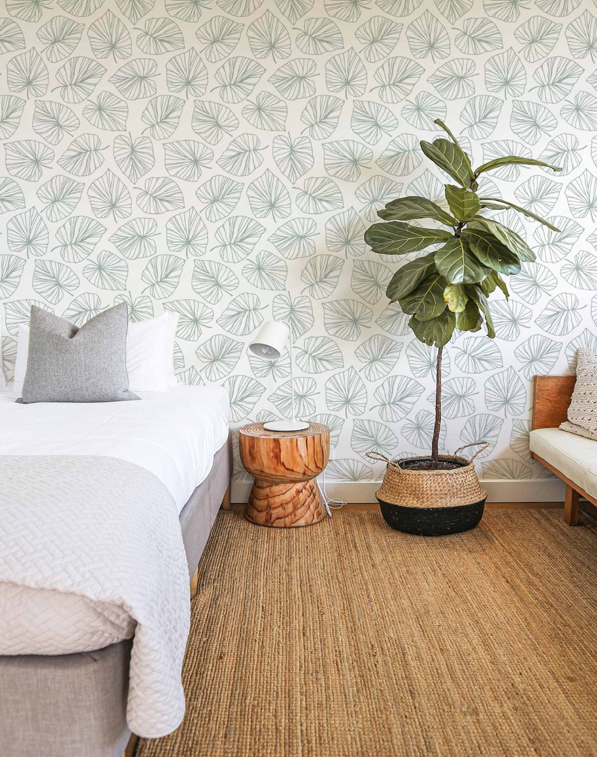 A cozy bedroom corner with the Line Art Tropical Leaf Wallpaper as a backdrop. The wallpaper features a simple line art design of large tropical leaves in sage green on a white background. The room includes a wooden side table, a white lamp, a bed with white and grey bedding, and a large potted plant, enhancing the natural and fresh atmosphere.