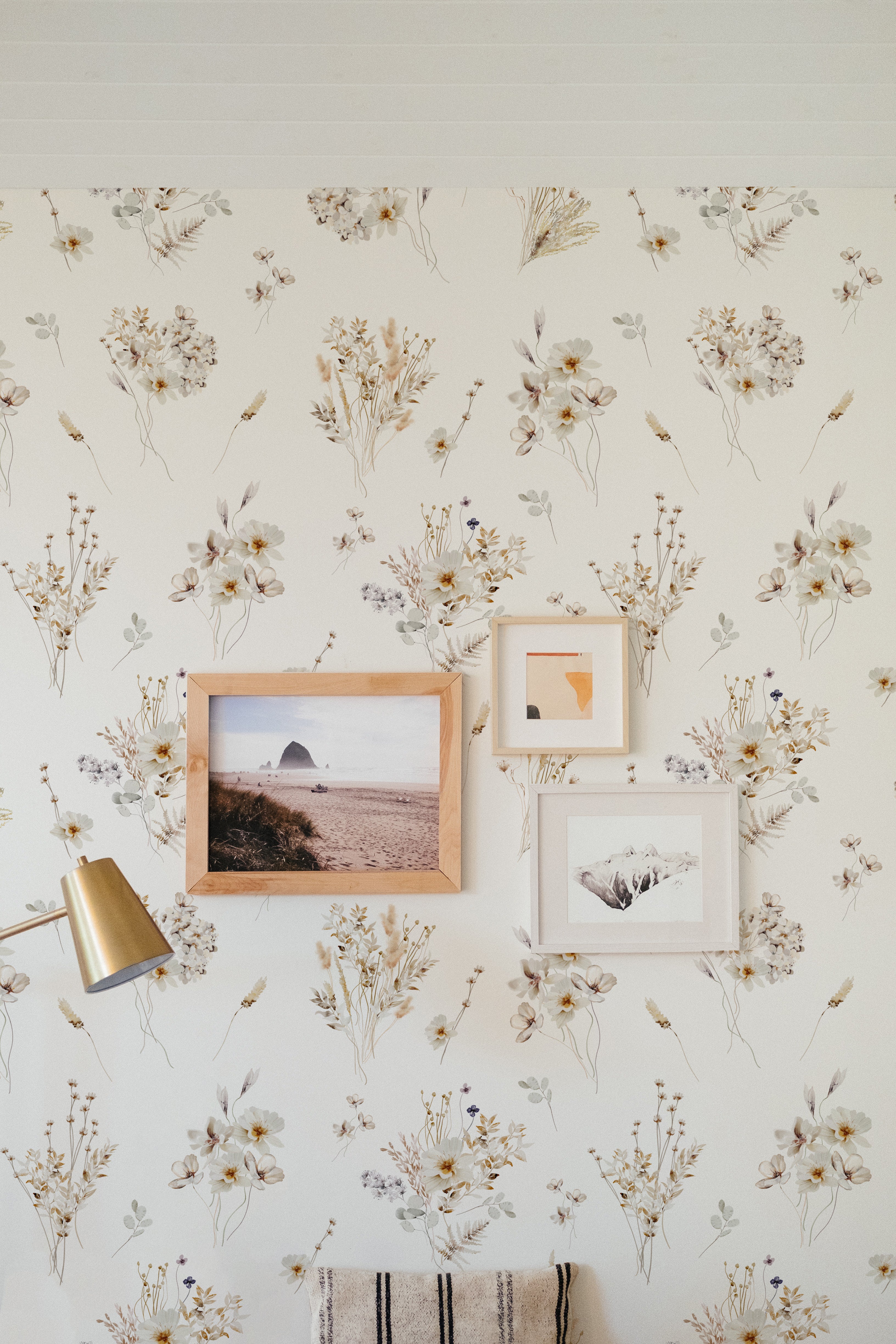 A charming interior wall adorned with 'Meadow Muse Wallpaper', interspersed with wooden frames of artwork, complemented by a vintage gold reading lamp, creating a cozy and artistic atmosphere.