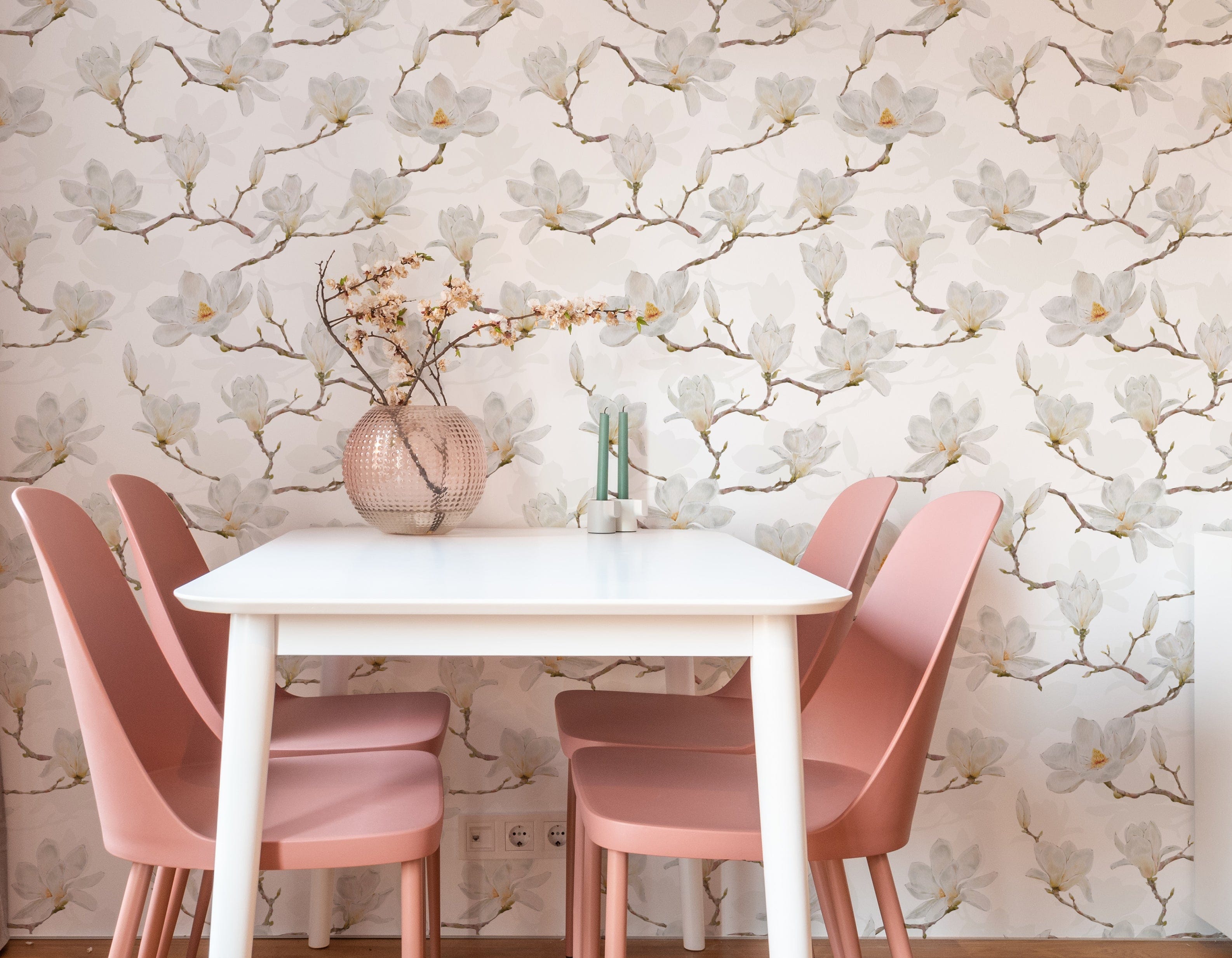 A minimalist dining area with a white table and pastel pink chairs. A textured glass vase with dried flowers sits in the center of the table, with the backdrop of white watercolour magnolia wallpaper enhancing the serene and stylish ambiance.