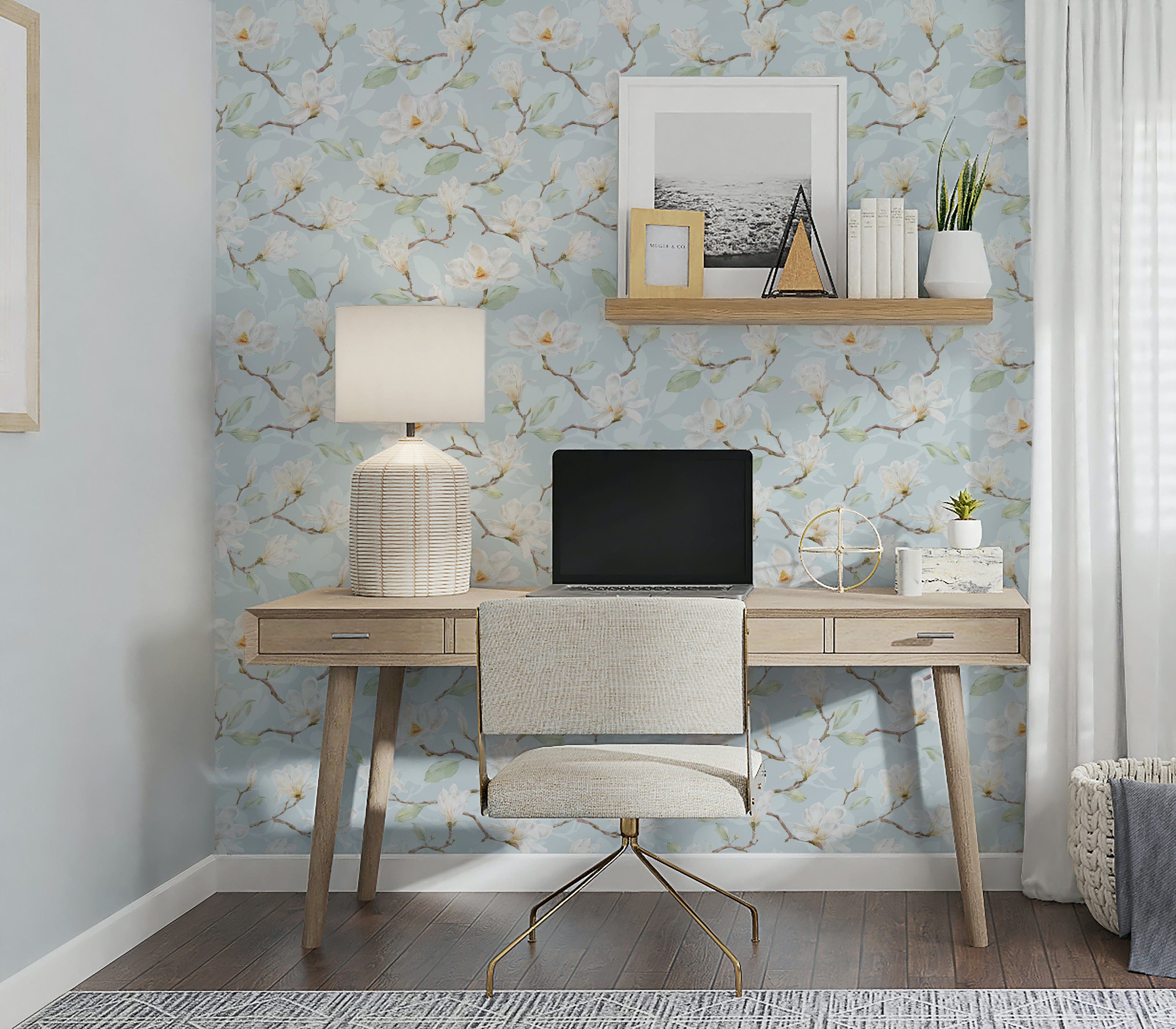 A home office setting with a wall adorned in 'Watercolour Magnolia Wallpaper II', showcasing light blue backdrop with delicate white and pale yellow magnolia blooms and soft green leaves. A wooden desk with a beige upholstered chair, topped with a cream lamp, gives the space a serene and creative atmosphere.