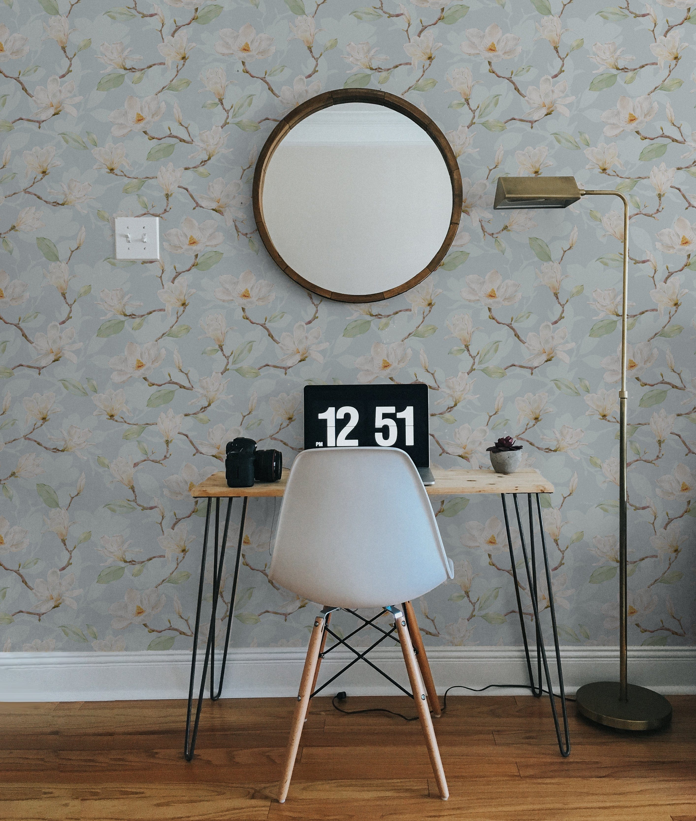 A stylish workspace featuring the 'Watercolour Magnolia Wallpaper II' on the wall, enhancing the area with its watercolor magnolias set against a pale blue background. A minimalist desk with a modern white chair and a sleek brass floor lamp complete the tranquil, inspired setting.