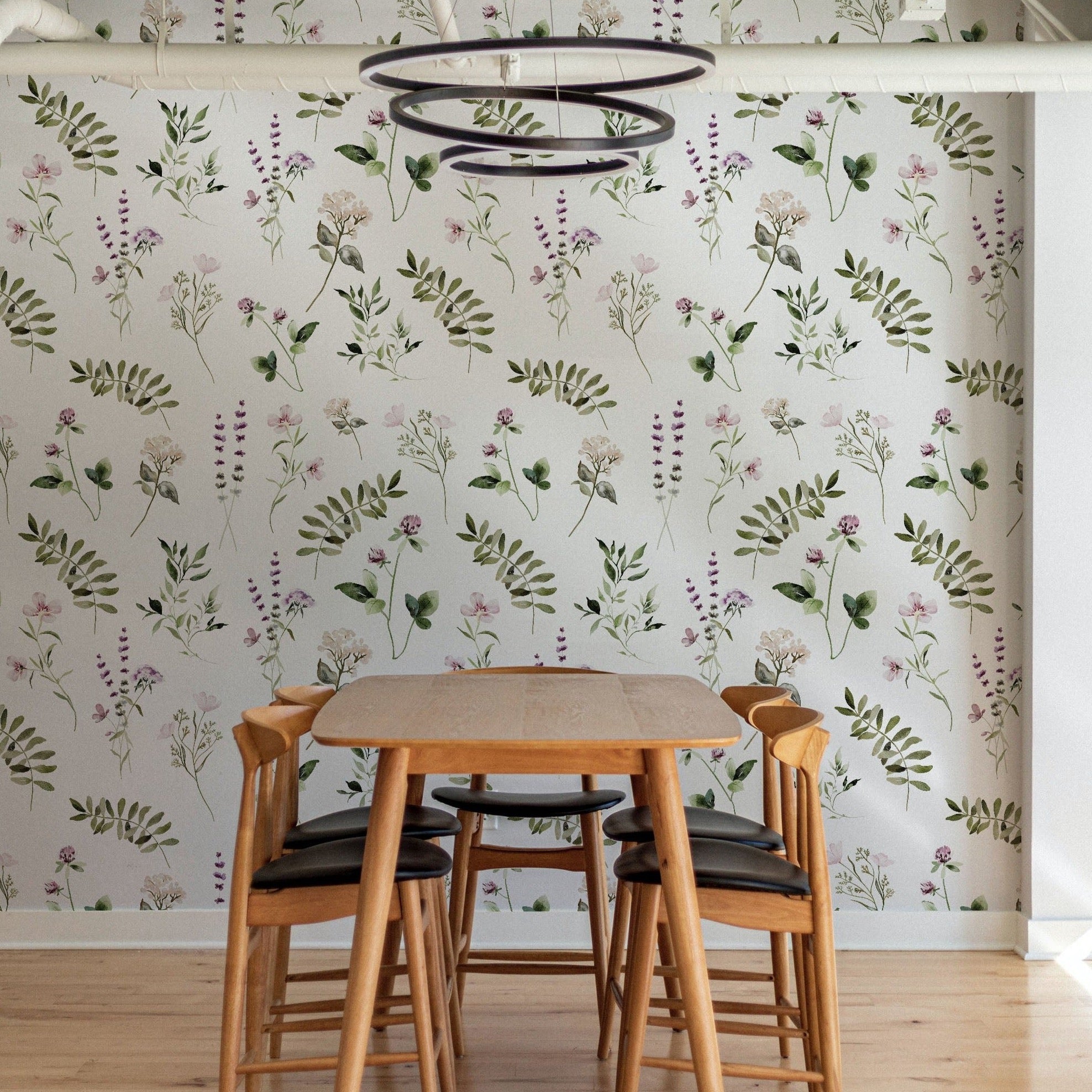 A dining area featuring walls covered with "Midsummer Watercolour Floral & Fern Wallpaper." The room is furnished with a simple wooden dining table and chairs, with the wallpaper providing a lively backdrop of green ferns and pastel flowers, creating a calming and inviting environment.