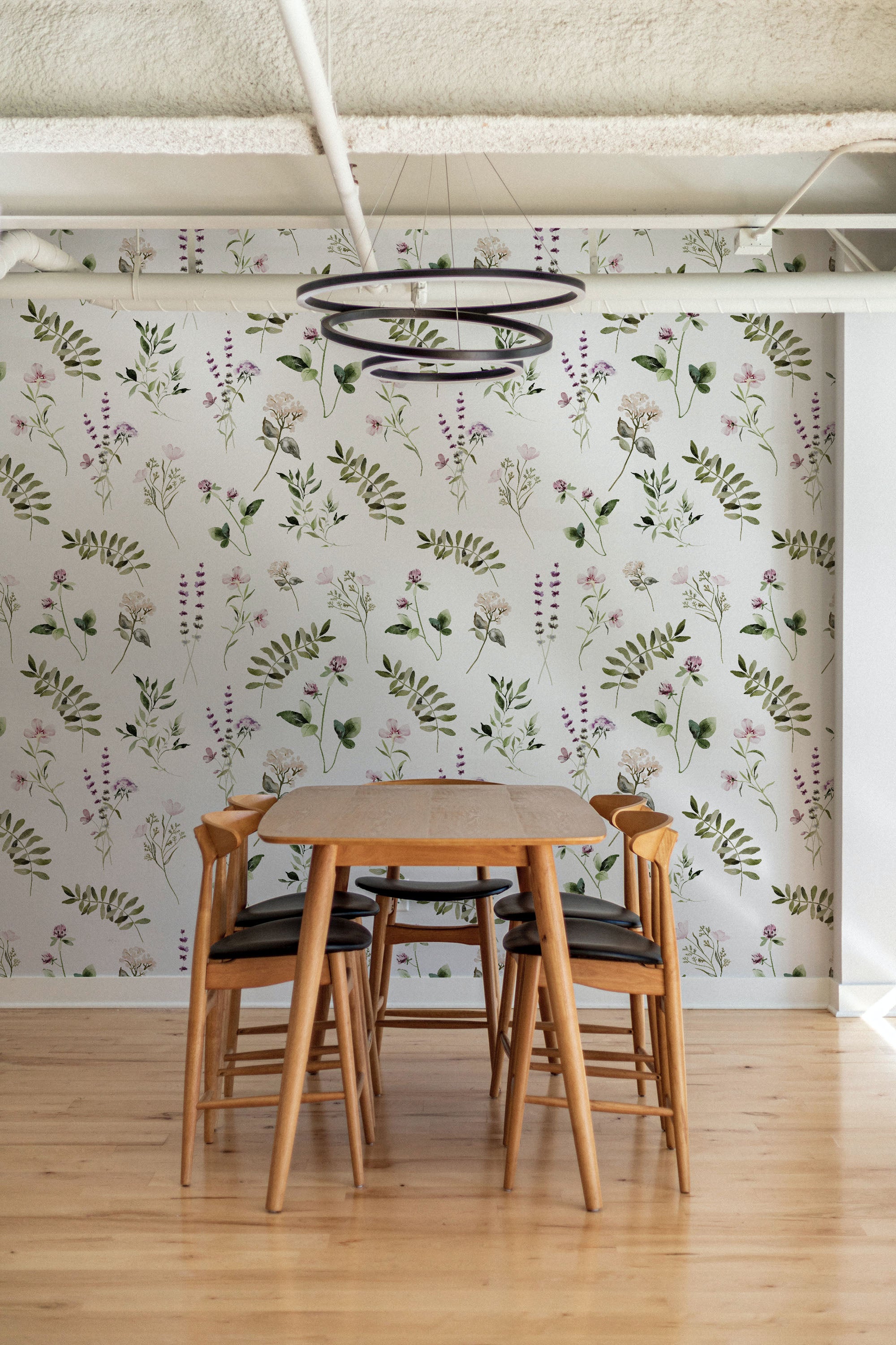A dining area featuring walls covered with "Midsummer Watercolour Floral & Fern Wallpaper." The room is furnished with a simple wooden dining table and chairs, with the wallpaper providing a lively backdrop of green ferns and pastel flowers, creating a calming and inviting environment.