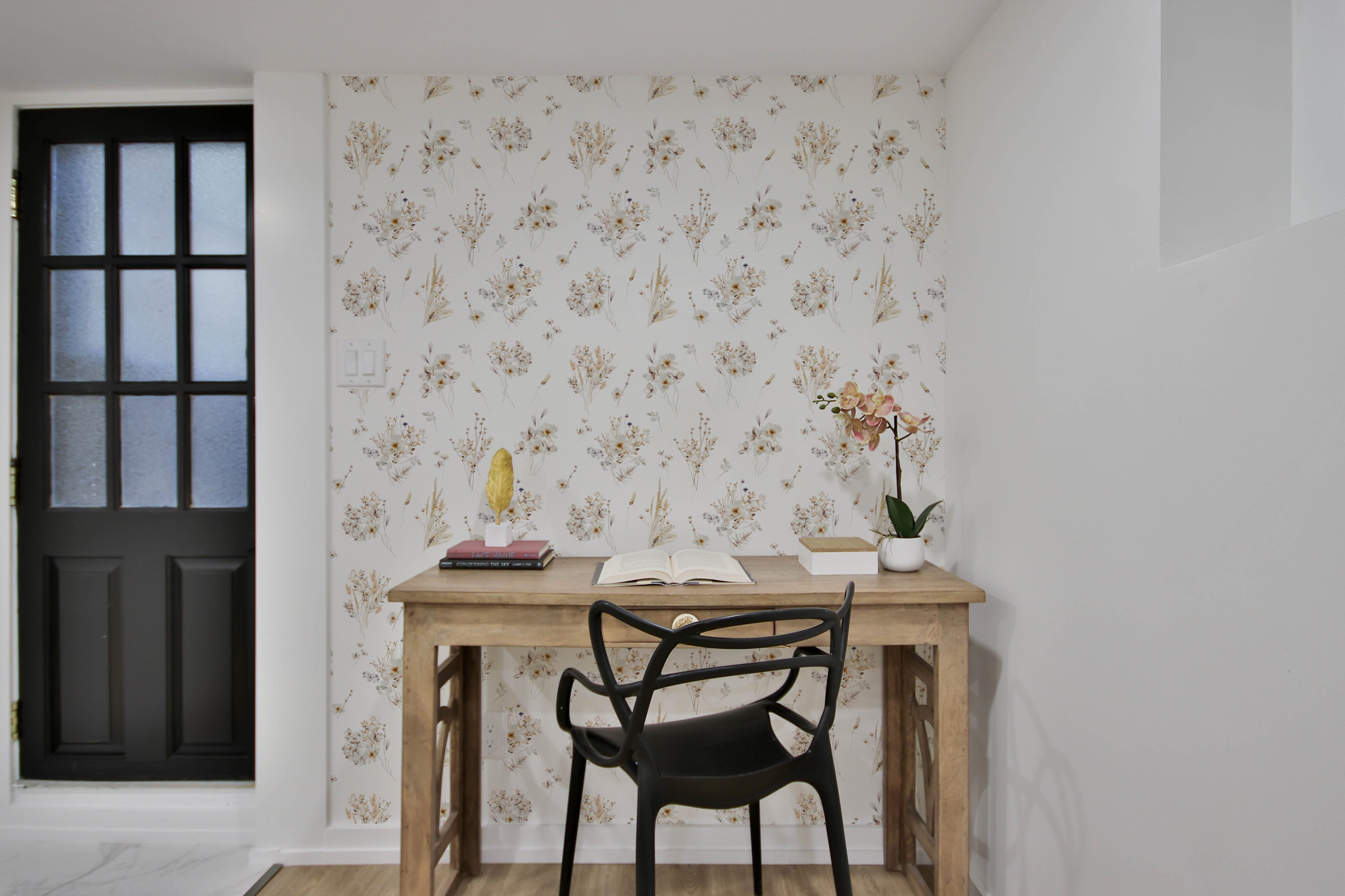 A modern workspace corner with the 'Meadow Muse Wallpaper' as a backdrop featuring a pattern of delicate dried flowers in soft brown and beige tones, complementing the sleek wooden desk and black chair.